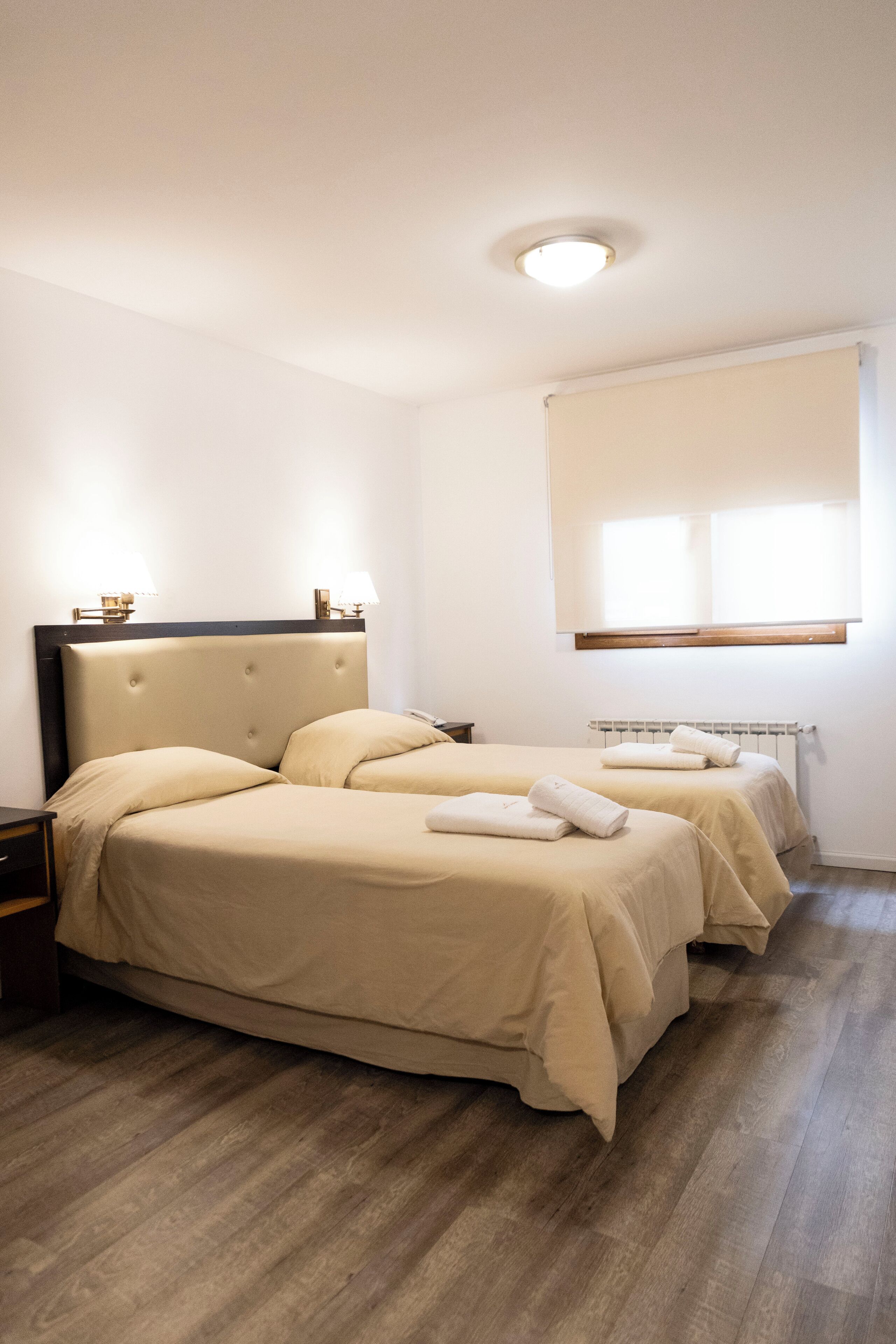 Een hotelkamer met twee bedden, een nachtkastje, een raam met zonwering en een radiator.