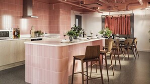 Une cuisine moderne avec un mur recouvert de carreaux roses, un îlot central doté de tabourets de bar et un coin repas agrémenté d’un rideau rouge en arrière-plan.