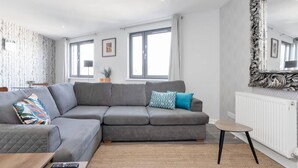 Ein modernes Wohnzimmer mit einem grauen Ecksofa, einem kleinen runden Tisch und einem großen, verzierten Spiegel.