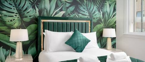 A bedroom with a green leaf-patterned wallpaper, a wooden headboard, white bedding, and two bedside lamps.