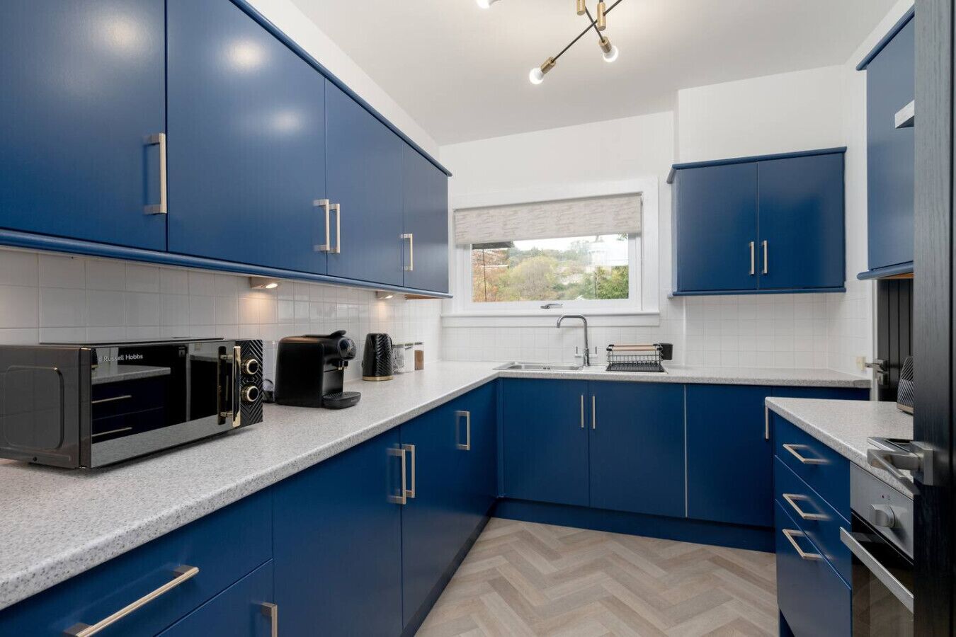 A modern kitchen with blue cabinets, a built-in microwave, a coffee maker, and a window with a view of greenery.
