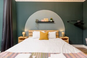 Une chambre à coucher avec un grand lit, deux tables de chevet, une arche murale incurvée et une étagère ornée d’objets décoratifs.