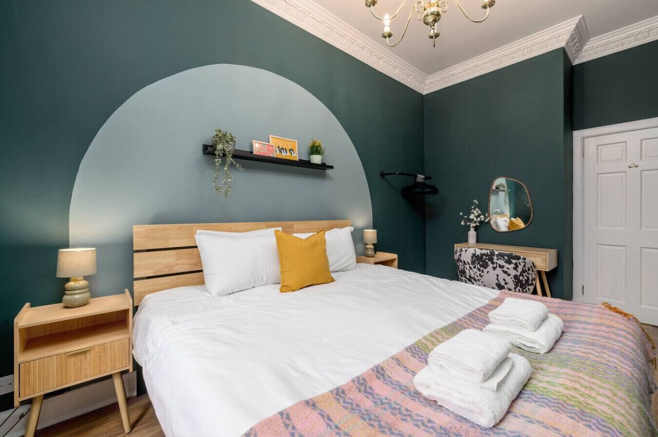 Quarto com cama, mesa de cabeceira, espelho e prateleira na parede com objetos decorativos.