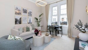 Ett modernt vardagsrum med en soffa, ett soffbord och en matplats. På väggen hänger inramade tavlor och det finns ett stort fönster med gardiner.