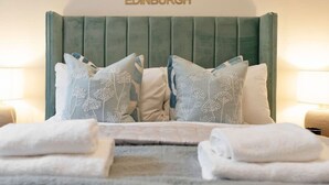 Ett hotellrum med en säng som har en grön, klädd sänggavel, flera kuddar med broderade blomstermotiv och prydligt vikta, vita handdukar på sängen.
