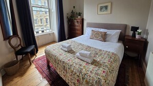 Quarto com cama, cômoda, cadeira, janela com cortinas e luminária.