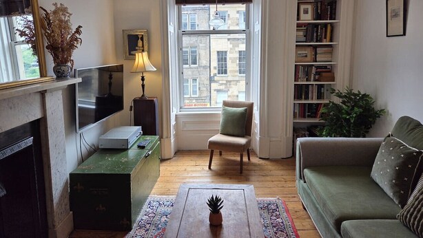 Uma sala de estar com lareira, estante de livros, sofá verde e janela com vista para edifícios.