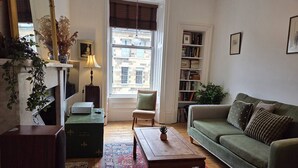 Uma sala de estar com um sofá verde, uma mesa de centro de madeira, um tapete estampado, uma estante de livros e uma janela com vista para edifícios.