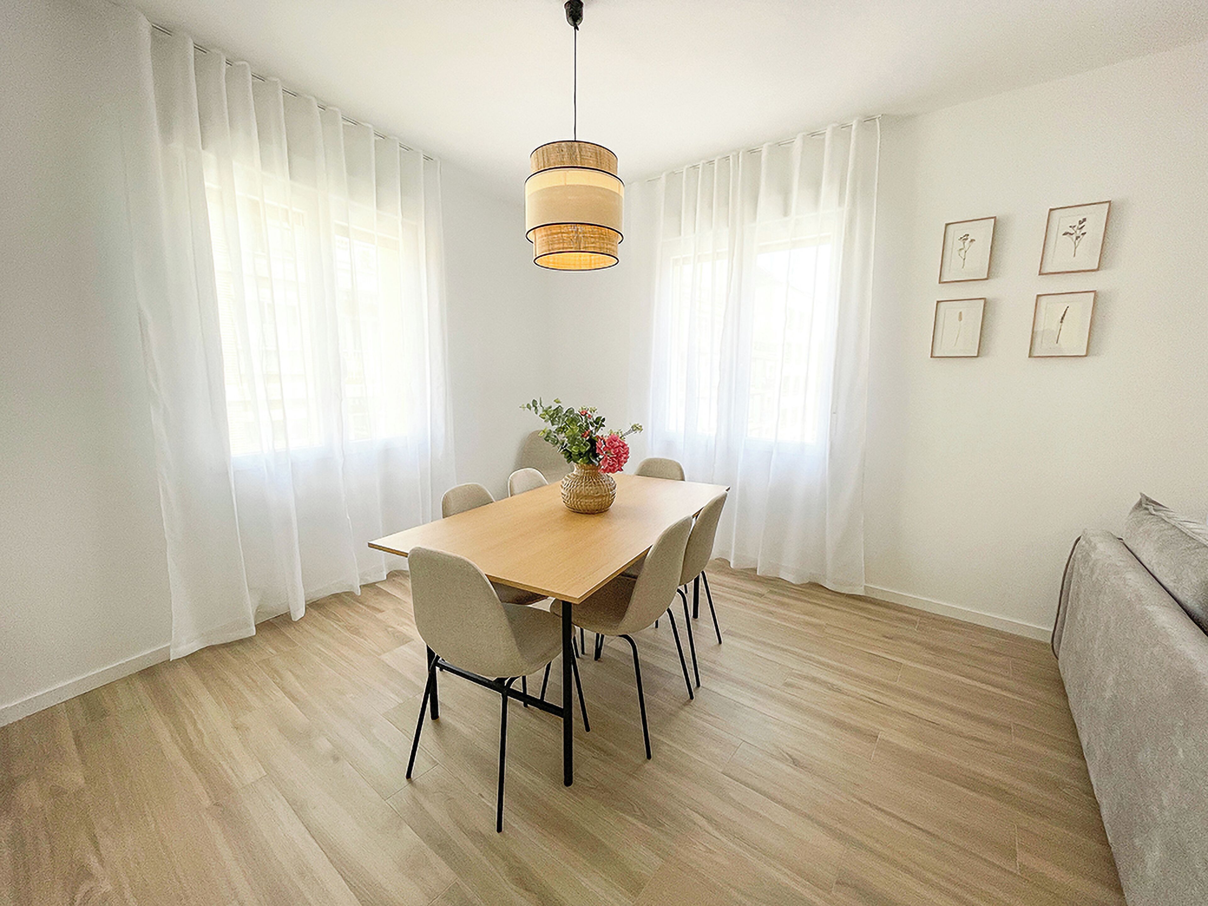 A dining area with a wooden table, six chairs, a vase with flowers, and a hanging light fixture.