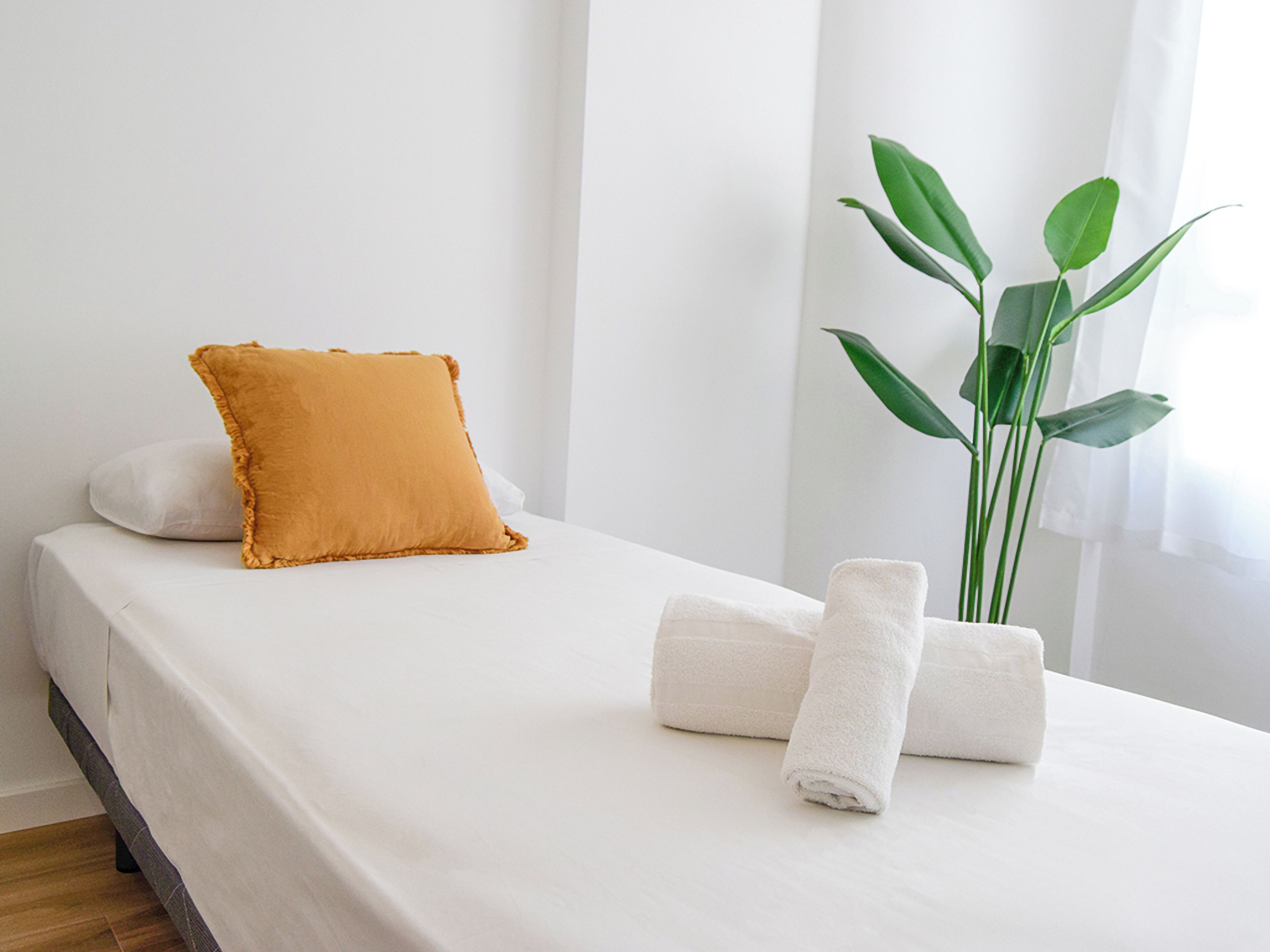 A neatly made bed with white bedding, a single orange pillow, and rolled white towels.