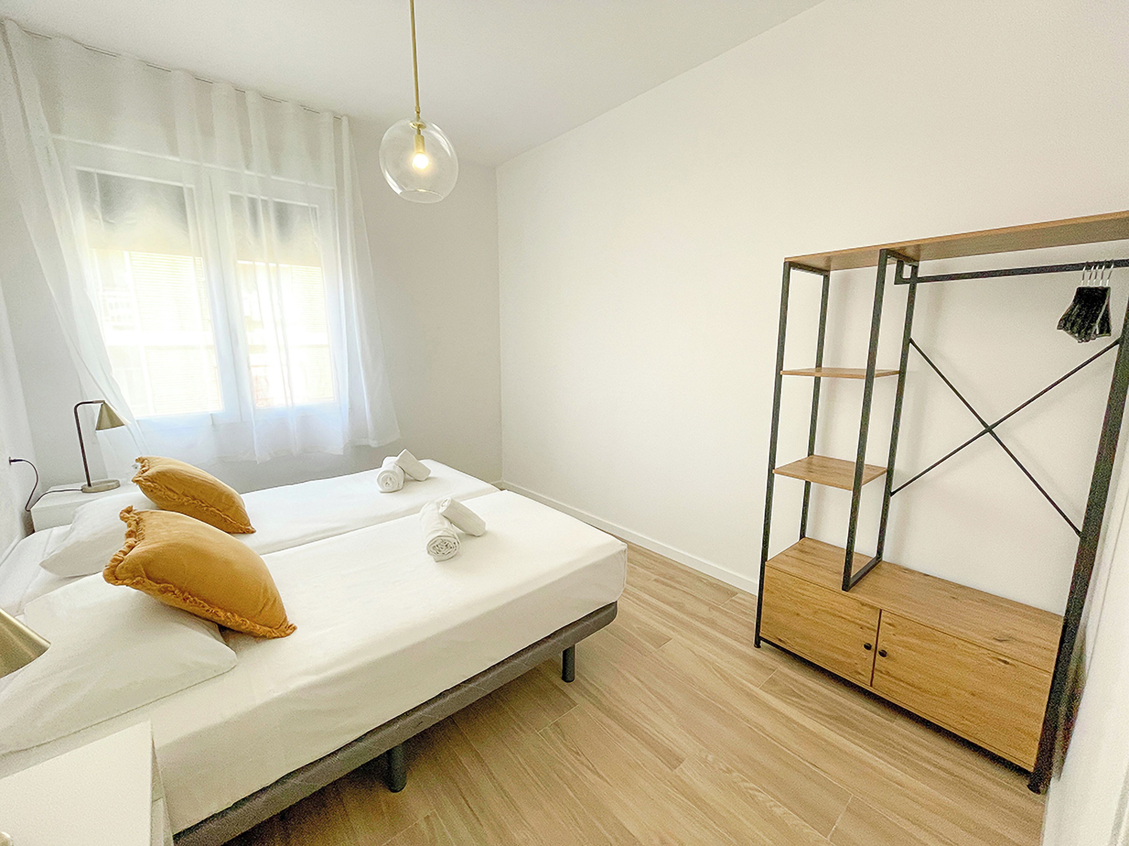 A bedroom with a bed, two pillows, a bedside table, a lamp, a wardrobe, and a hanging light fixture.