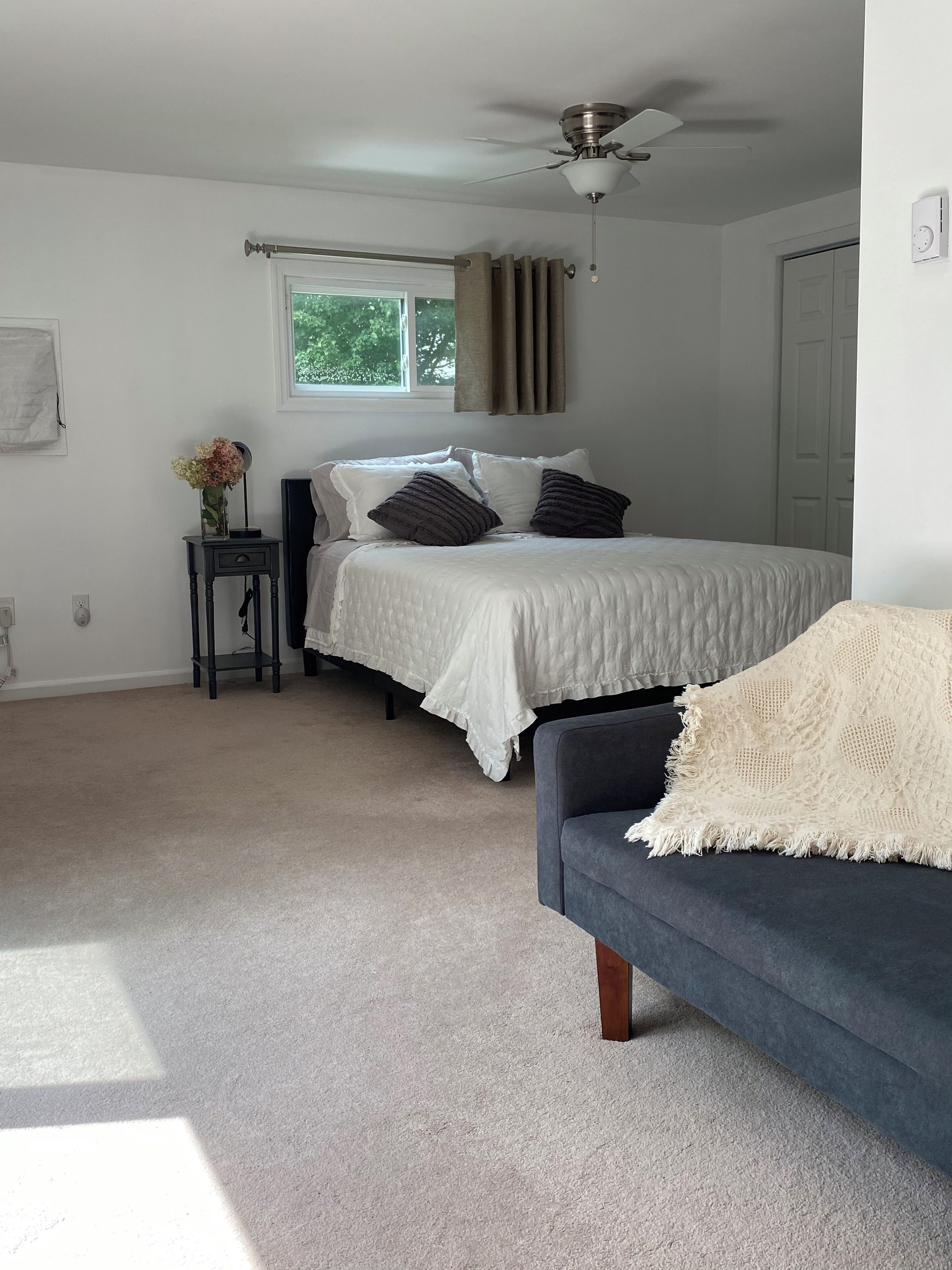 A bedroom with a bed, a bedside table with a vase, a ceiling fan, and a window with curtains.
