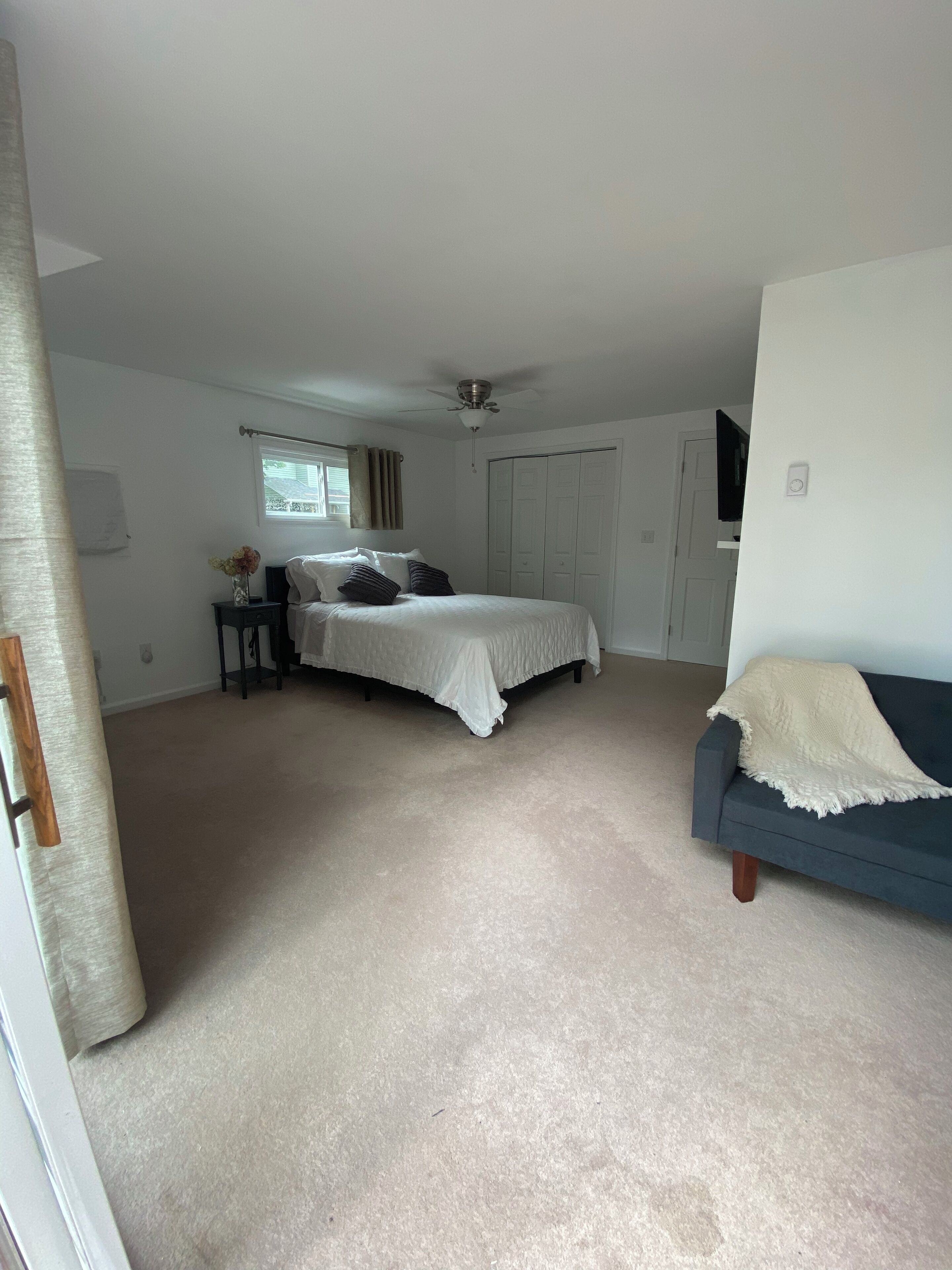 Ein Schlafzimmer mit einem Bett, einem Nachttisch, einer Kommode, einem Deckenventilator, einem Fenster mit Vorhängen und einem Sofa.