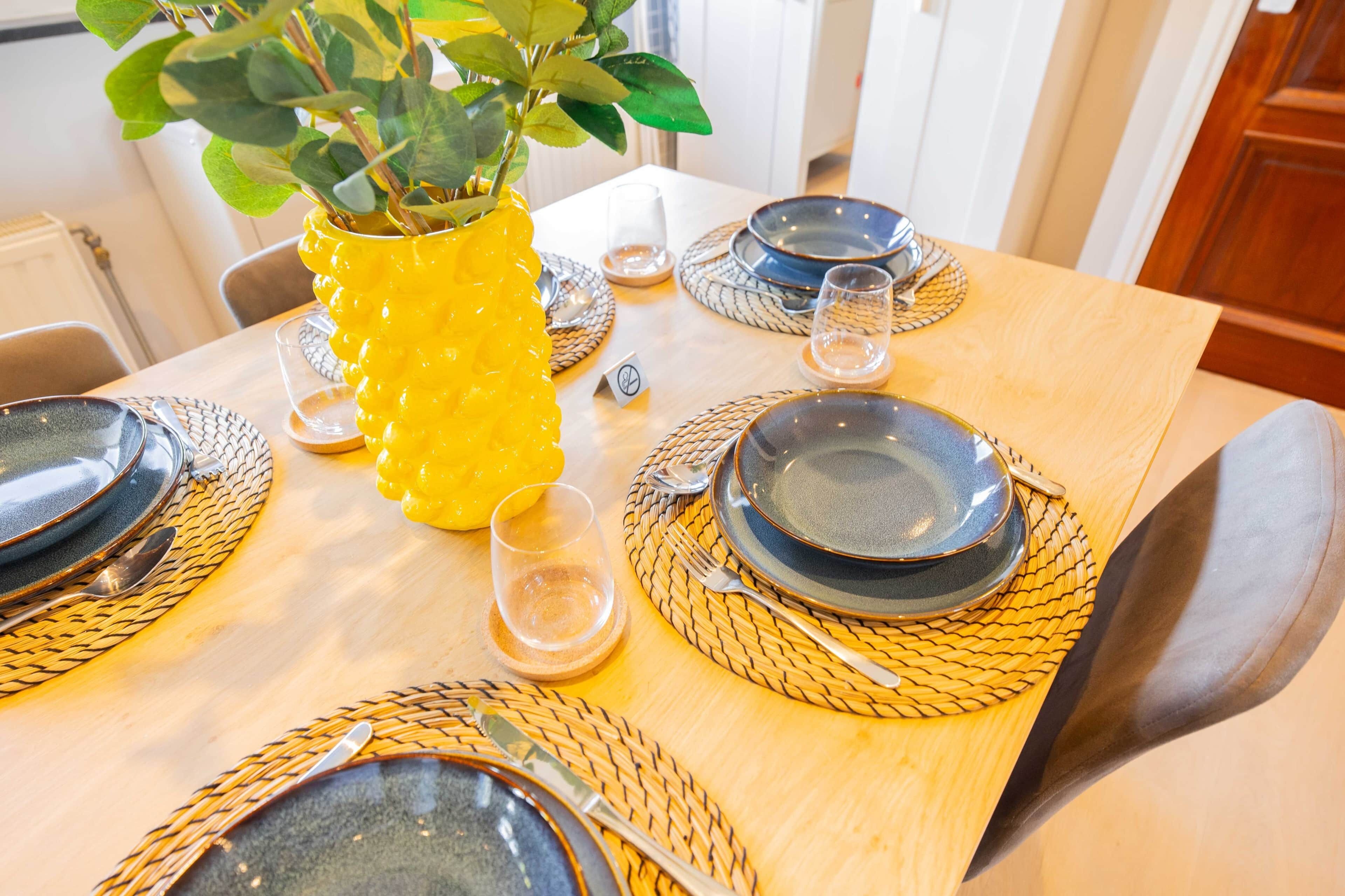 Una mesa de comedor dispuesta con platos, cubiertos y un jarrón amarillo con hojas verdes.