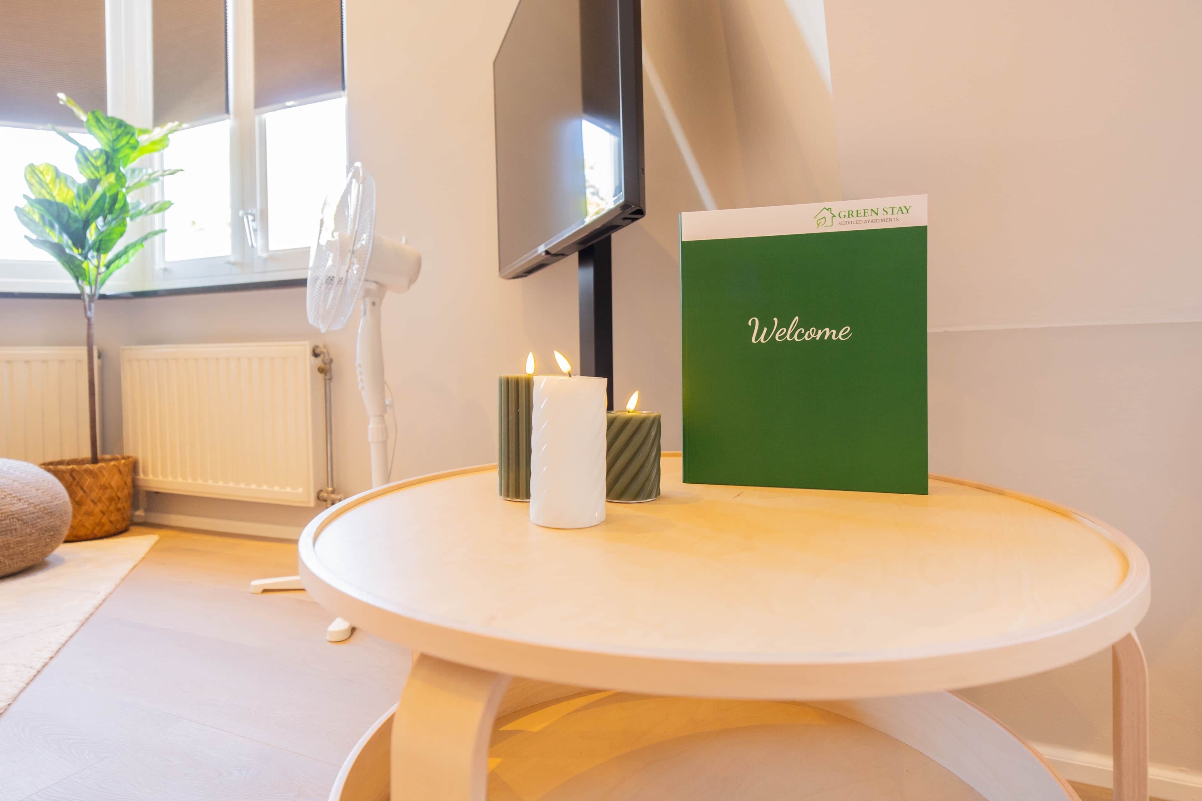 Een kamer met een ronde tafel, een groen welkomstbord, een witte kaars en een plant in pot.