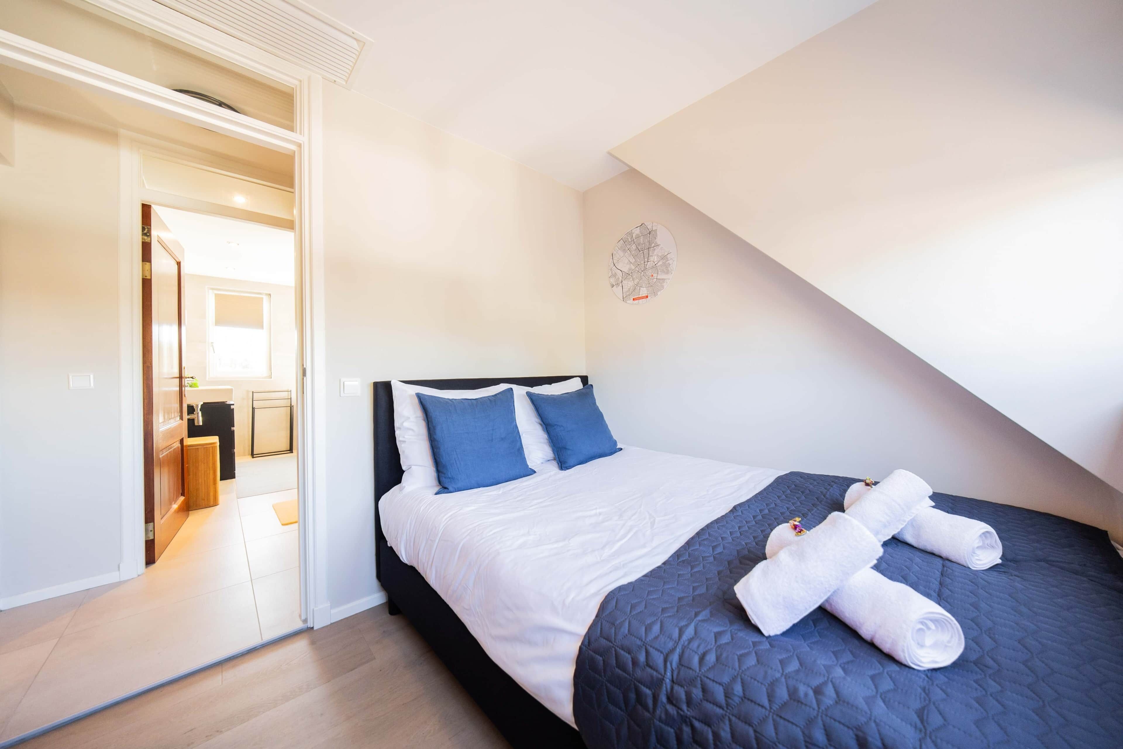A bedroom with a bed, pillows, a quilted blanket, and rolled towels.