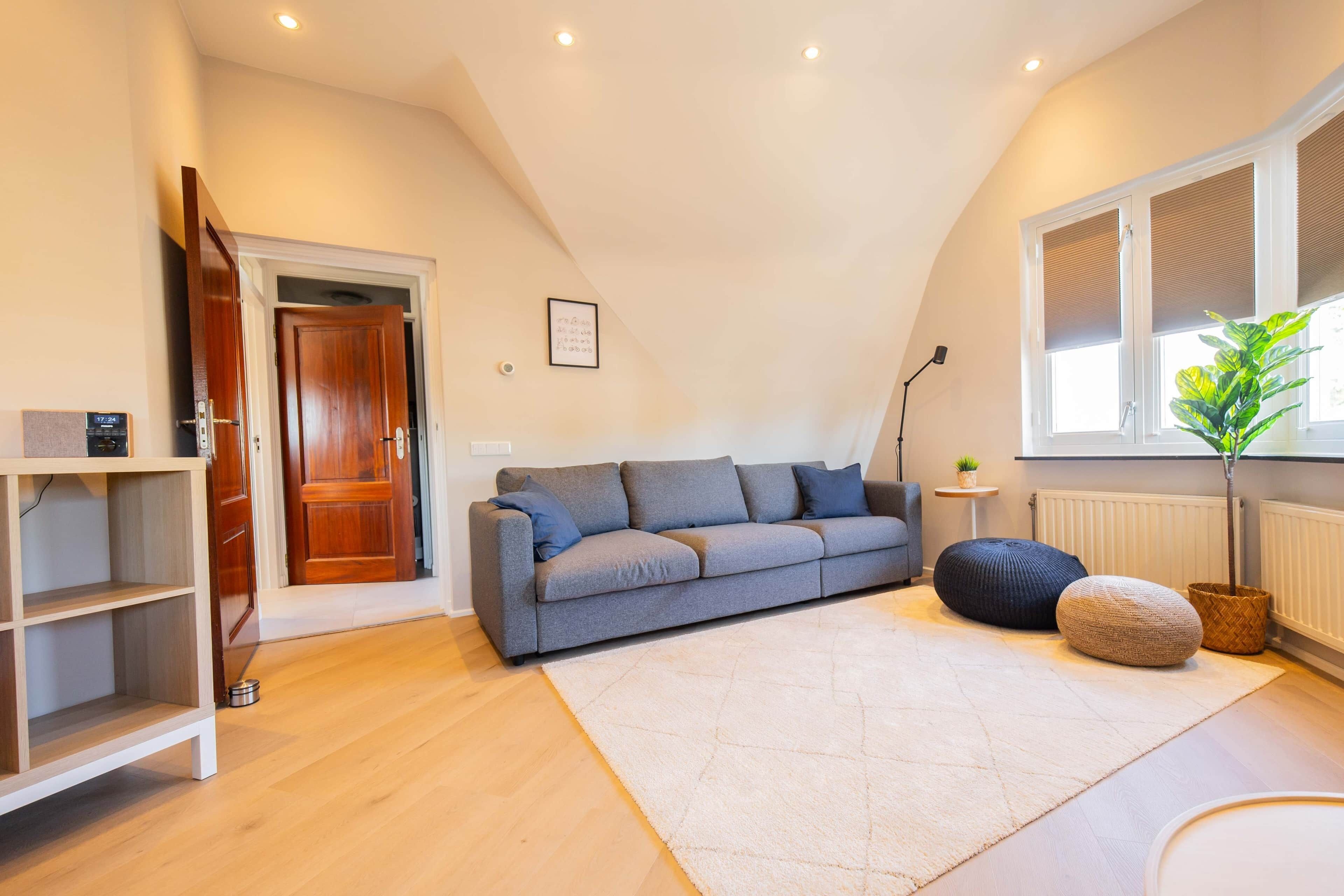 A modern living room with a grey sofa, a white rug, wooden flooring, and a window with blinds.