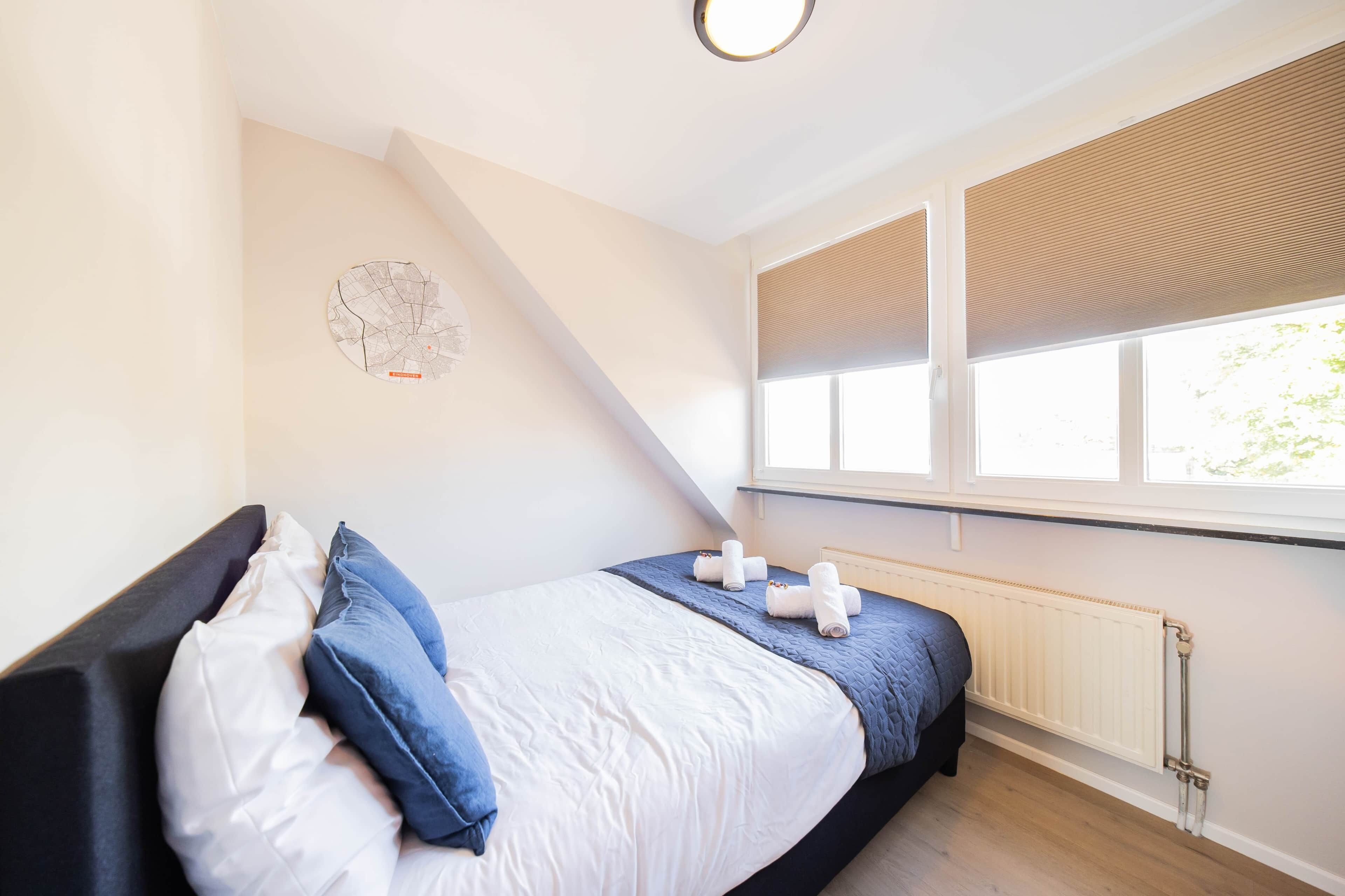 Un dormitorio con cama, almohadas, una ventana con persianas, un radiador y un reloj en la pared.