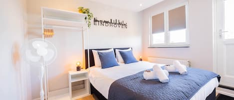 A hotel room with a bed, bedside tables, a wall-mounted fan, and a window with blinds.