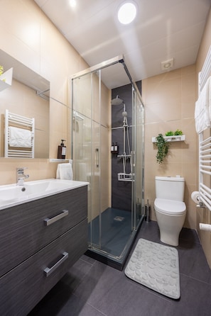 Ein modernes Badezimmer mit einer Duschkabine aus Glas, einem weißen Waschbecken, einem dunklen Schminktisch und einer Toilette.
