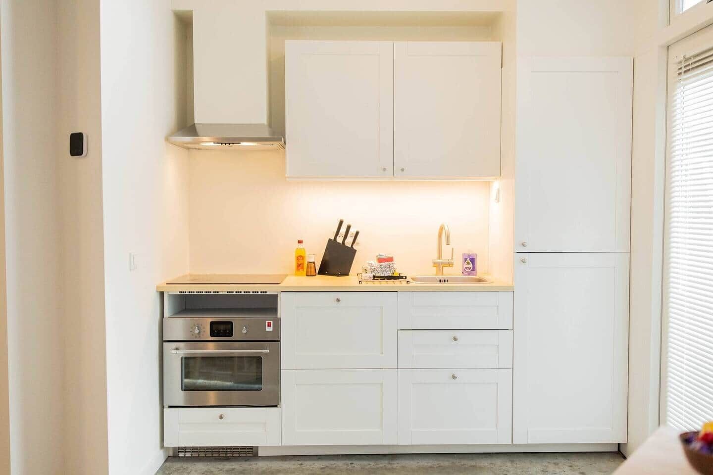 Een compacte keuken met witte kastjes, een roestvrijstalen oven, een inbouwmagnetron en een klein aanrecht met een messenblok en enkele spullen.