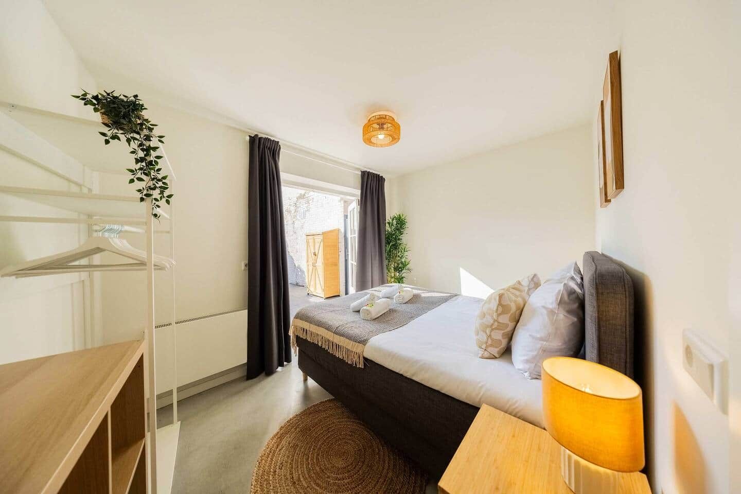 Een hotelkamer met een bed, nachtkastje, lamp en uitzicht op buiten.
