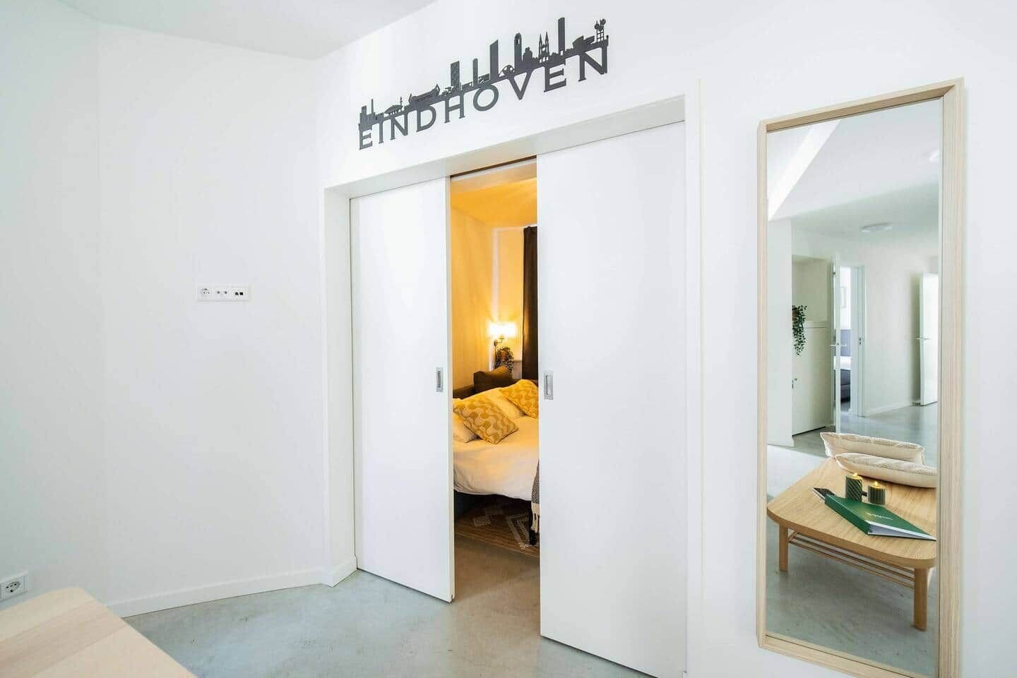 A hotel room with a bed, a bedside table, a mirror, and a door leading to another room.