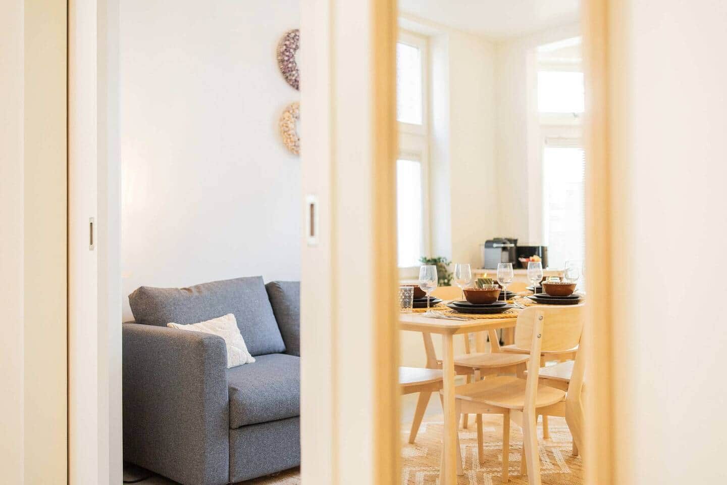 Un comedor con una mesa dispuesta para cuatro personas, un sofá gris y un ventanal con luz natural.