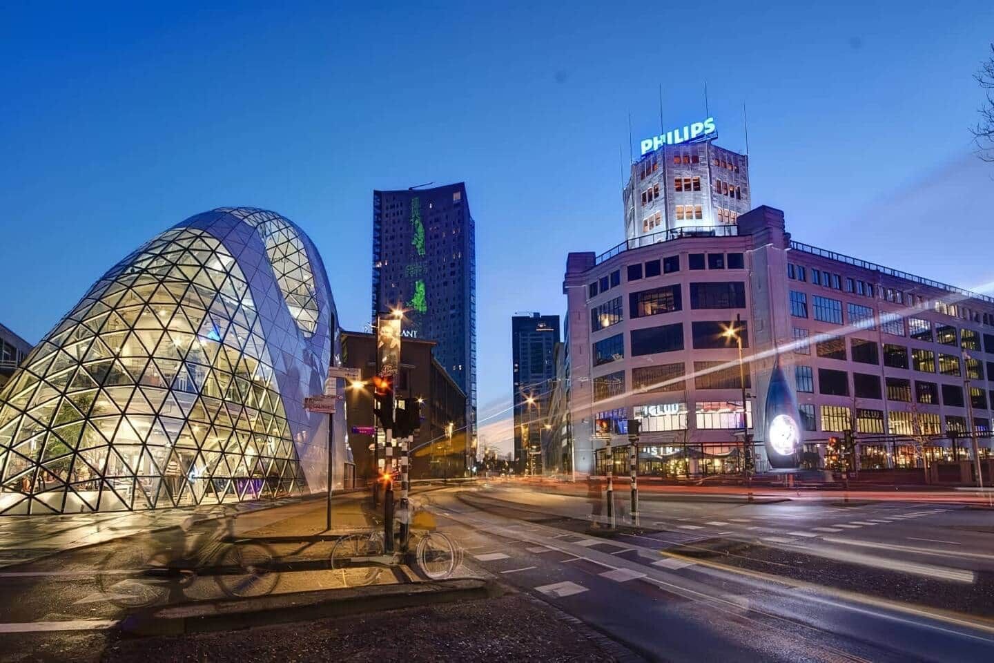 En modern stadsbild med en markant, geodätisk kupolkonstruktion, en hög byggnad med en Philips-skylt och en flervåningsbyggnad med upplysta skyltar.