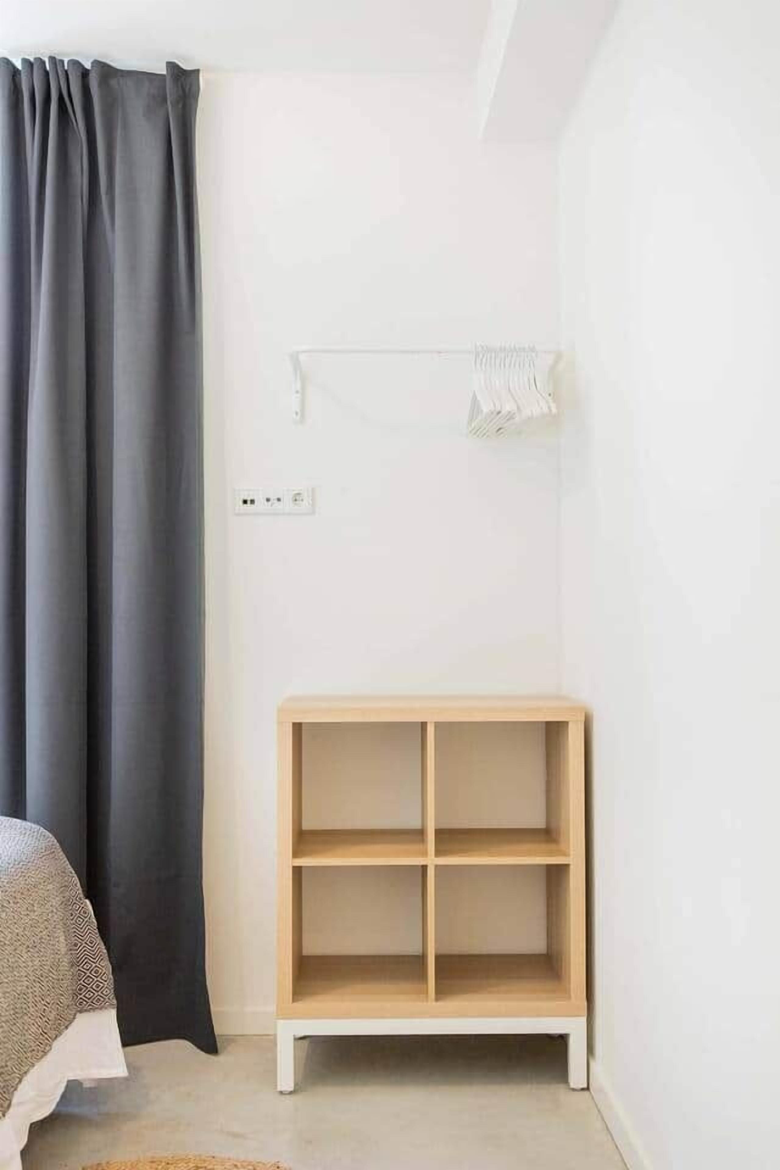 Een slaapkamer met een bed, een donker gordijn, een witte muur, een houten wandplank en een witte hanglamp.