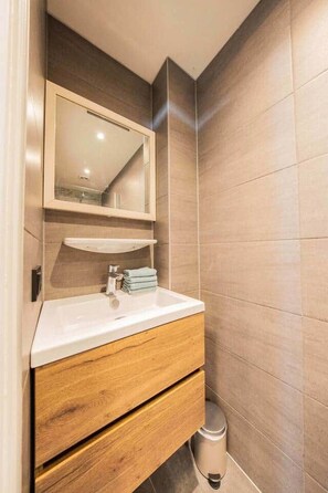 Une salle de bain moderne avec un lavabo blanc, un meuble-lavabo en bois, un miroir et un mur recouvert de carreaux.