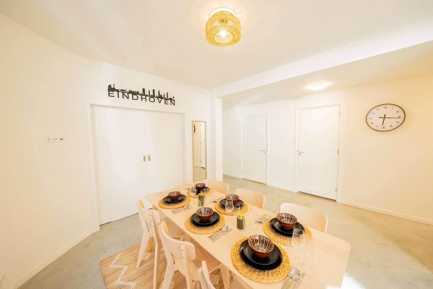 Een eetkamer met een gedekte tafel voor vier personen, witte wanden, een klok en een bord met een stadsgezicht.