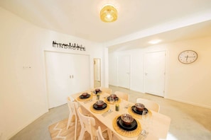 Une salle à manger avec une table dressée pour quatre personnes, des murs blancs, une horloge et un tableau représentant un paysage urbain.
