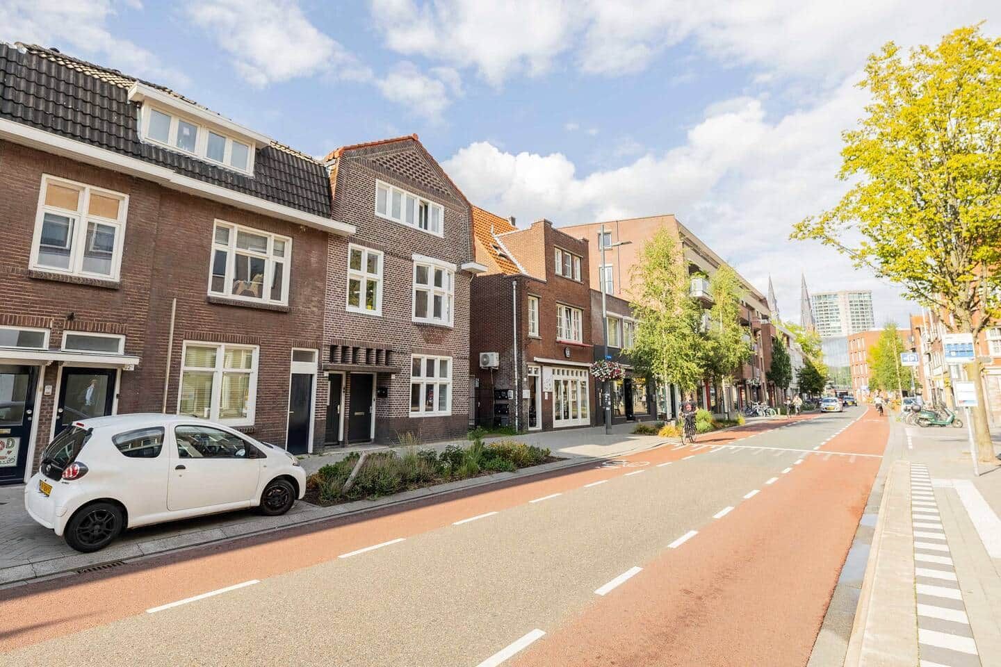Une rue bordée d’immeubles en briques, une voiture blanche garée sur le bord et un cycliste sur la route.