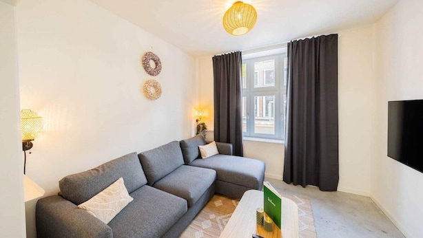 Un salon moderne avec un canapé d’angle gris, un téléviseur à écran plat fixé au mur et une petite table basse en bois.