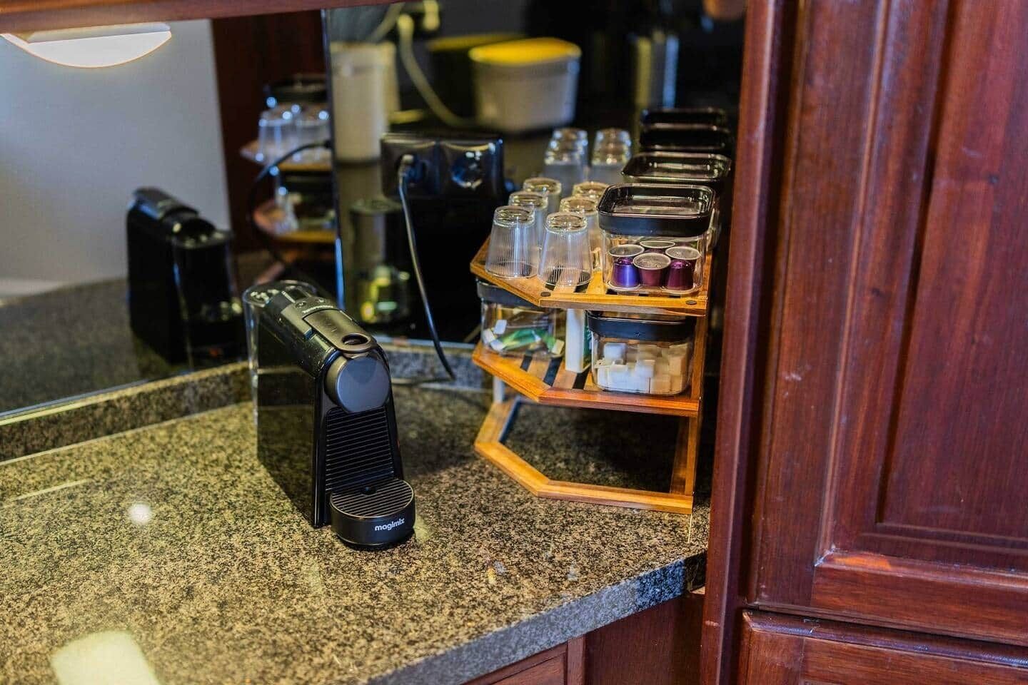 Une machine à café posée sur un plan de travail en granit, avec une étagère en bois où sont rangés des bocaux et des récipients.
