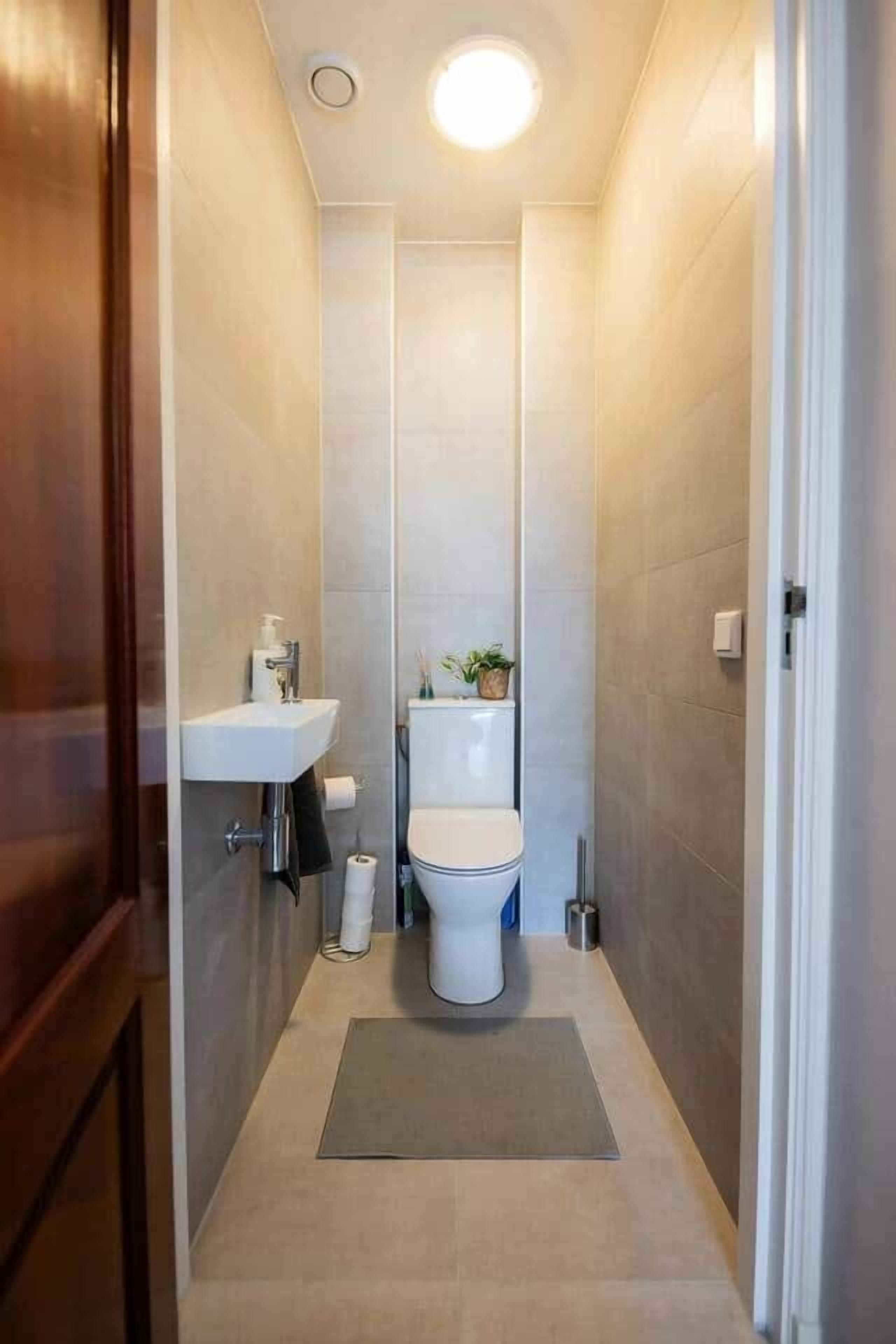 A narrow bathroom with a toilet, sink, and a small mat on the floor.
