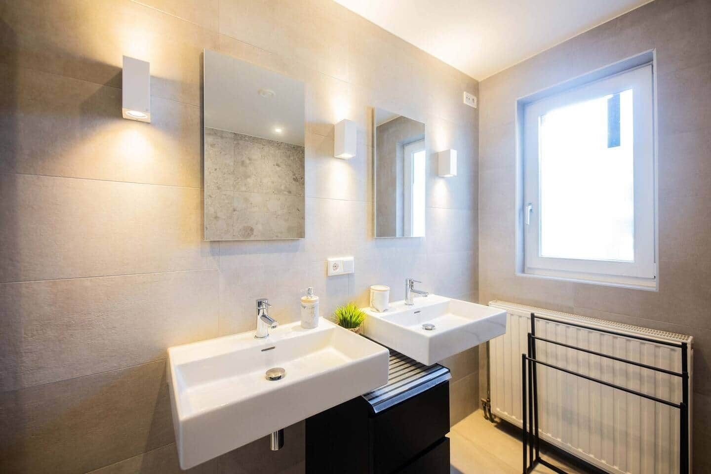 A modern bathroom with two sinks, a large mirror, and a window providing natural light.