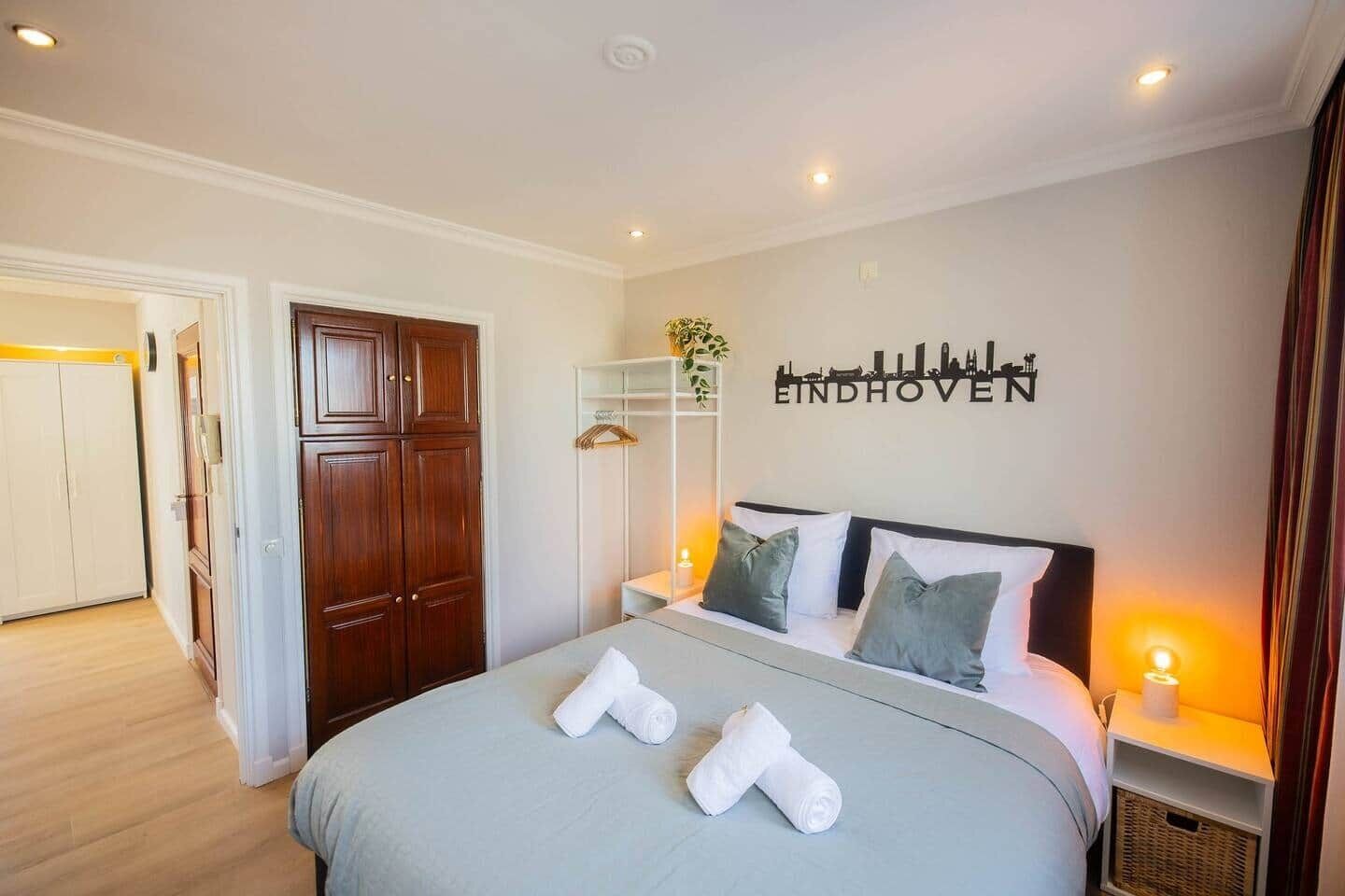A hotel room with a bed, a wardrobe, a wardrobe mirror, a shelf with a plant, and a wall with the text "EINDHOVEN".