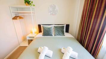 Hotellihuone, jossa on sänky, kaksi tyynyä, yöpöytä lampun kanssa, amppelissa roikkuva kasvi ja seinäkoriste.