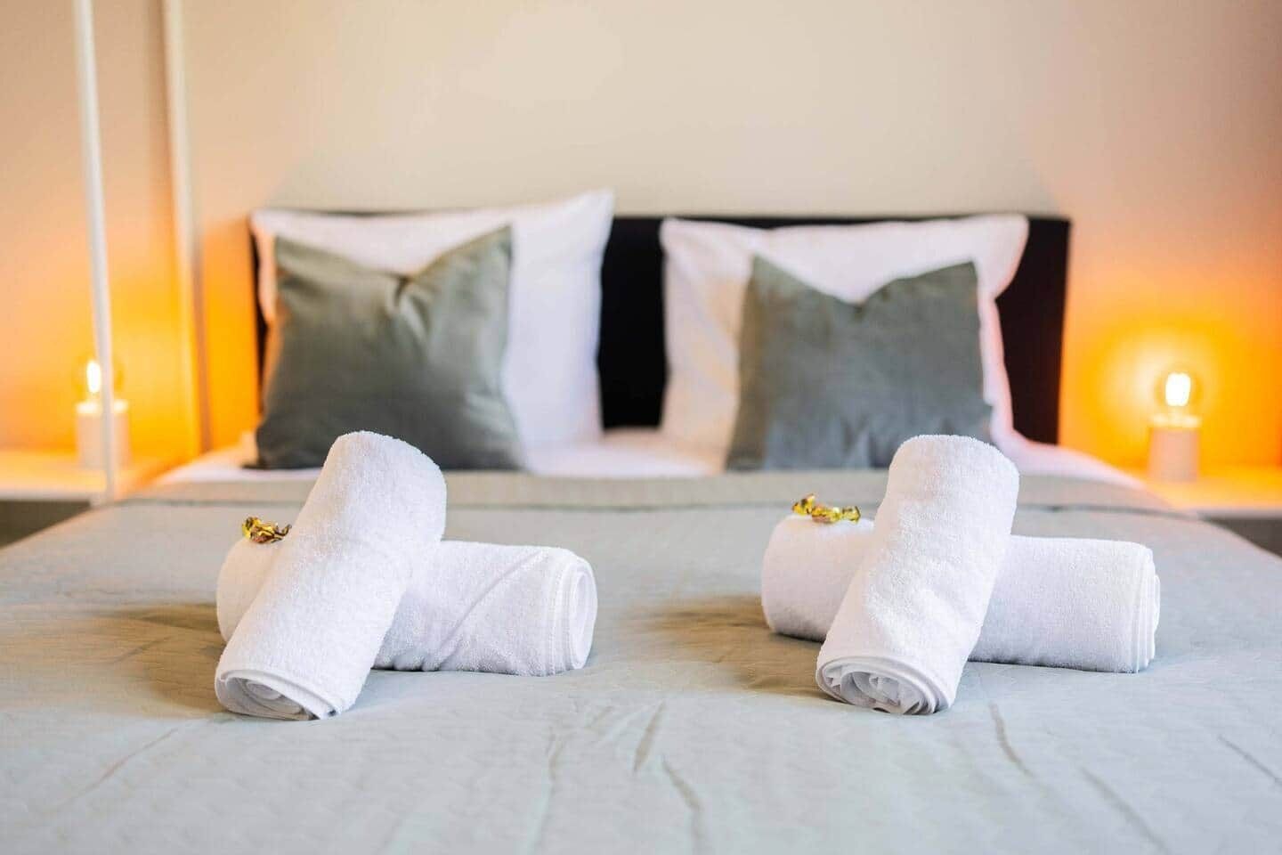 A neatly made bed with two rolled towels and a pillow, a bedside table with a candle, and a nightstand with a lamp.