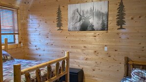 Interior de cabaña con dos camas de madera, un ventilador de techo y un cuadro de un paisaje forestal.