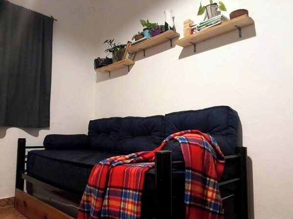 Un sofá oscuro con una manta escocesa, un estante de madera con plantas y libros, y una cortina negra.