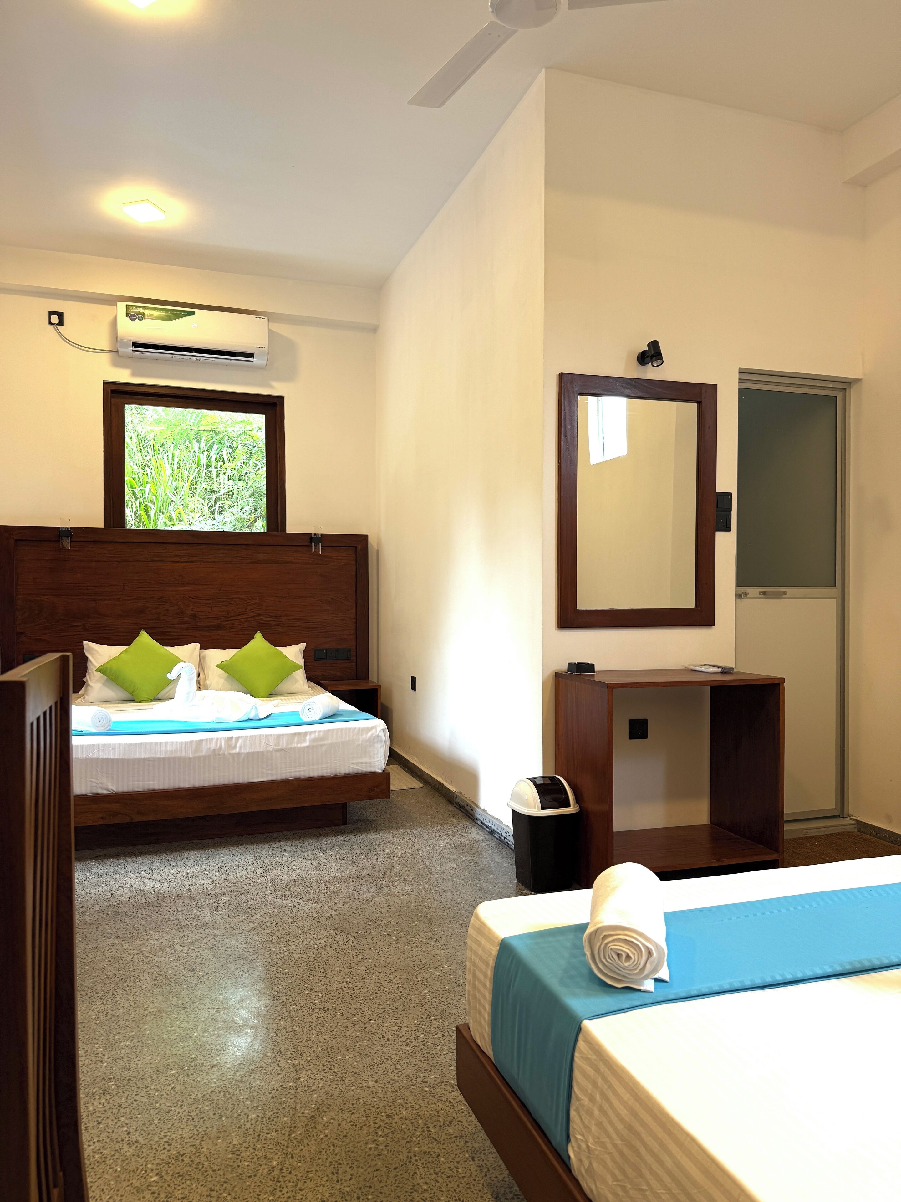 A hotel room with two beds, a window with a view of greenery, an air conditioning unit, a mirror, and a small wooden cabinet.