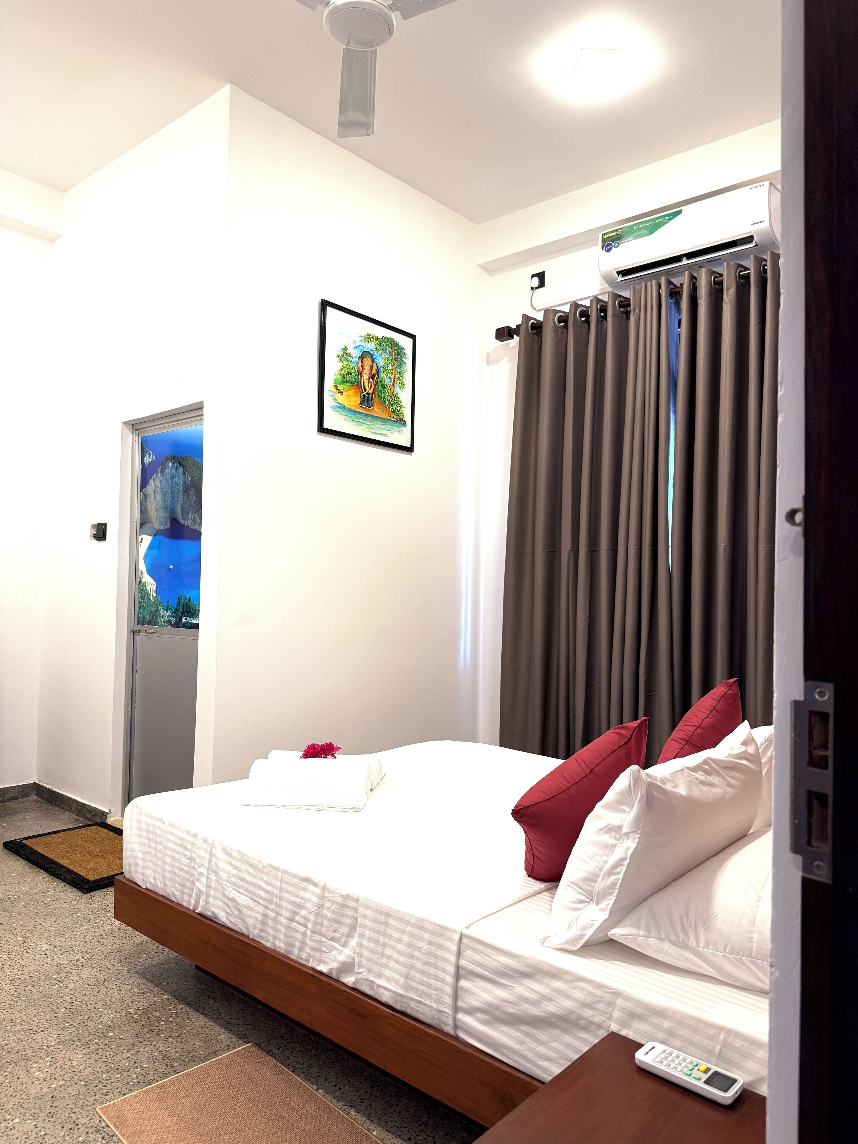A hotel room with a bed, a remote control, a picture on the wall, and a door leading to another room.