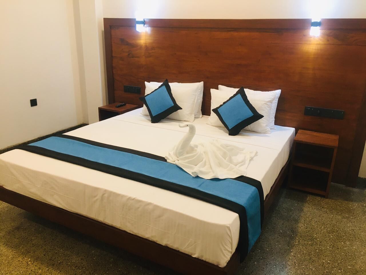 Una cama bien tendida con una colcha azul y blanca, dos almohadas y una almohada con forma de cisne. Se observa un cabecero de madera y una mesita de noche con un control remoto.