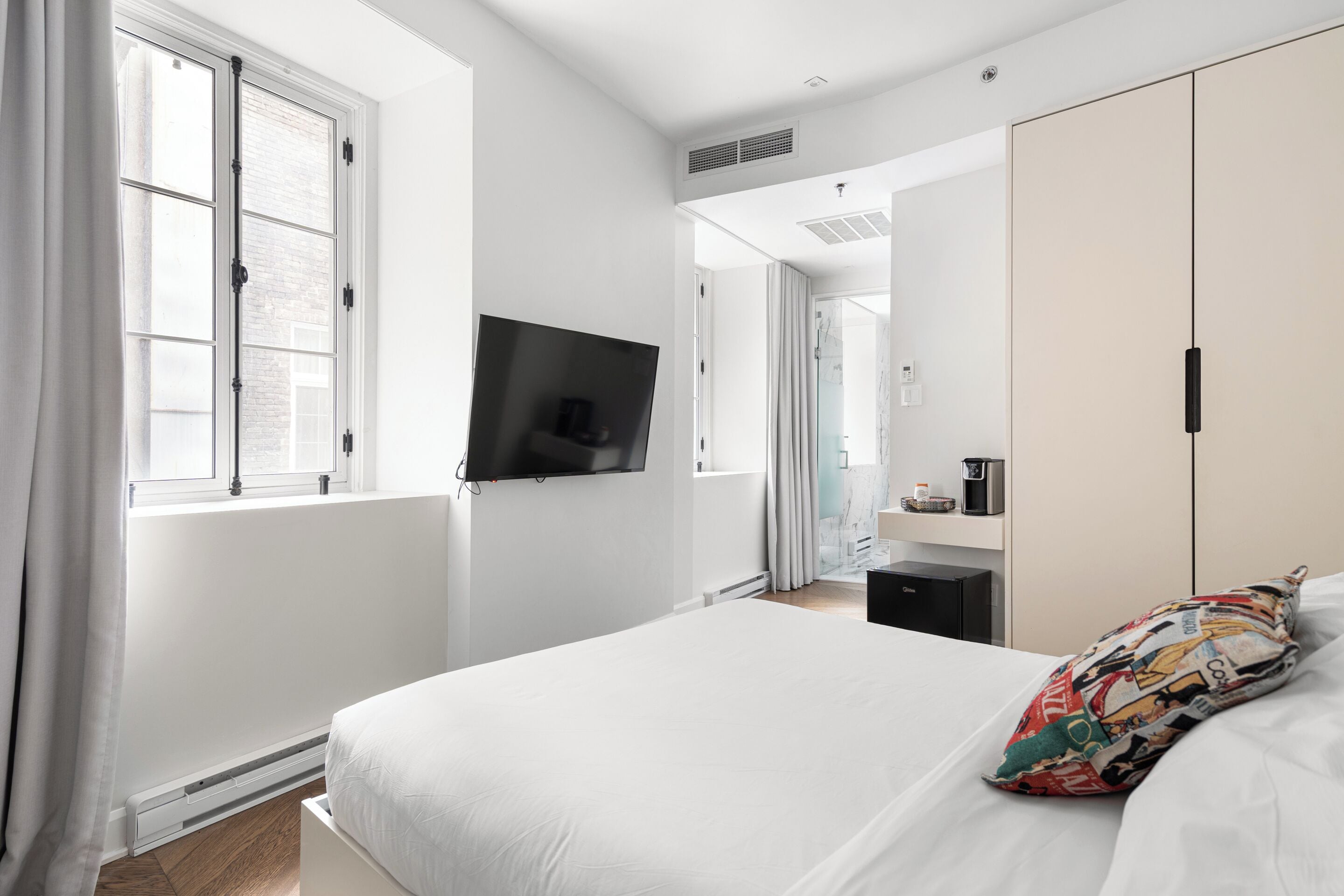 Un dormitorio moderno con una cama grande, un televisor de pantalla plana montado en la pared, un armario y un pequeño calentador debajo de la ventana.