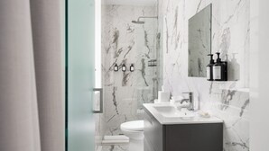 A modern bathroom with a marble shower, a toilet, and a vanity with a mirror and toiletries. - The Bowery Lofts Hotel (Montréal)