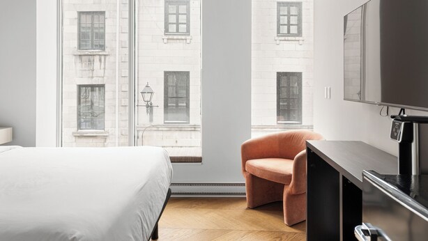 A modern hotel room with a large bed, a seating area with a chair, a flat-screen TV, and a view of a building through the window. - The Bowery Lofts Hotel (Montréal)