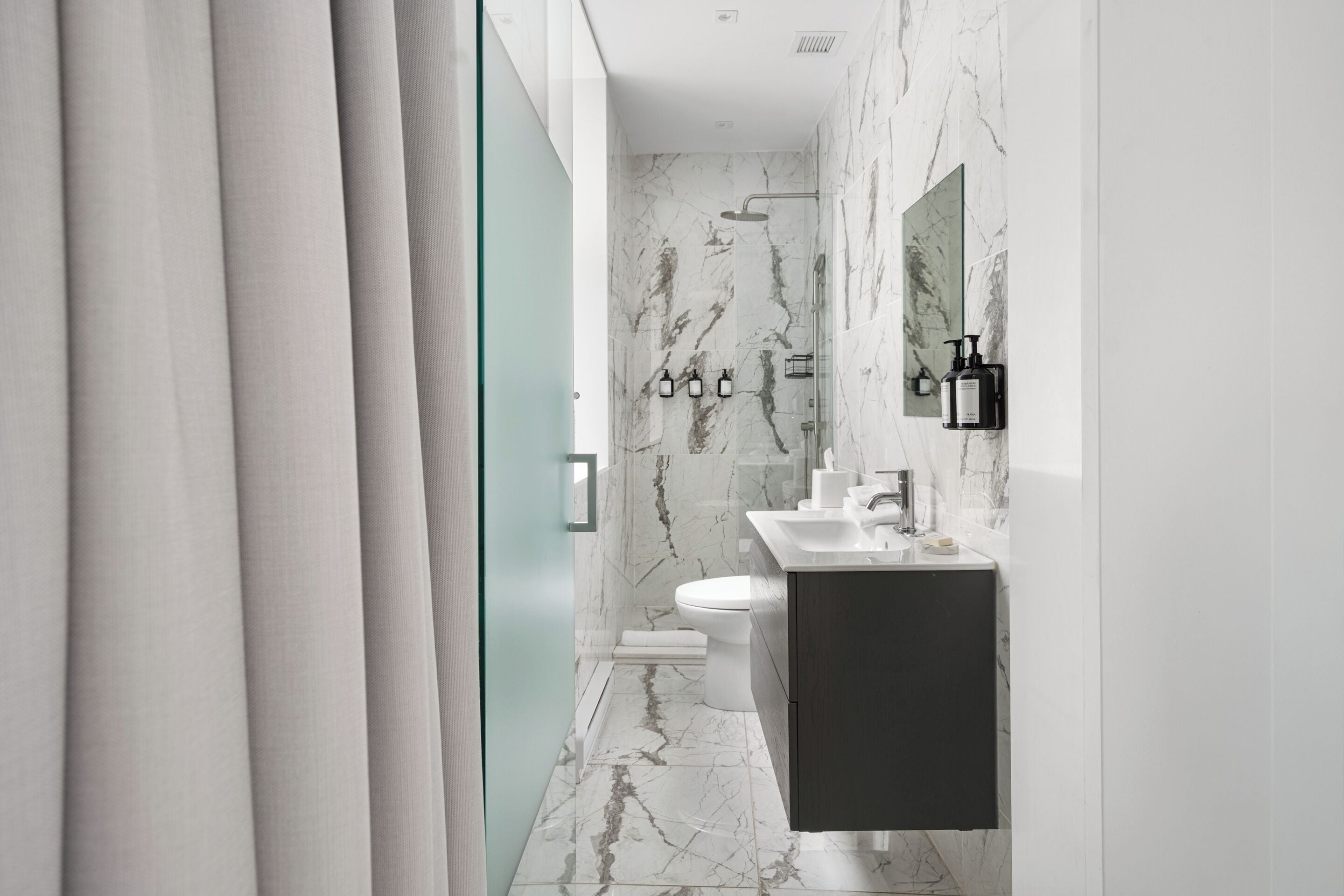 A modern bathroom with a marble floor, a white toilet, a dark vanity, and a glass shower enclosure.